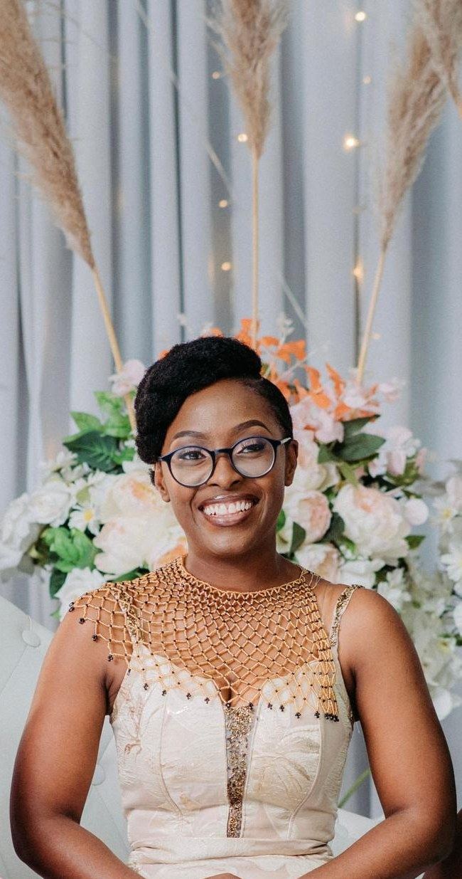 Danielle smiling, wearing round glasses and a beaded gold collar, in front of white flowers and pampas grass