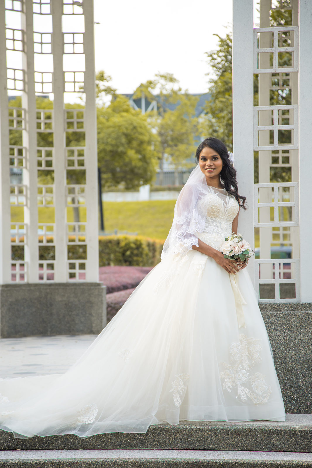 Aghalya in a white lace gown beneath a white pergola