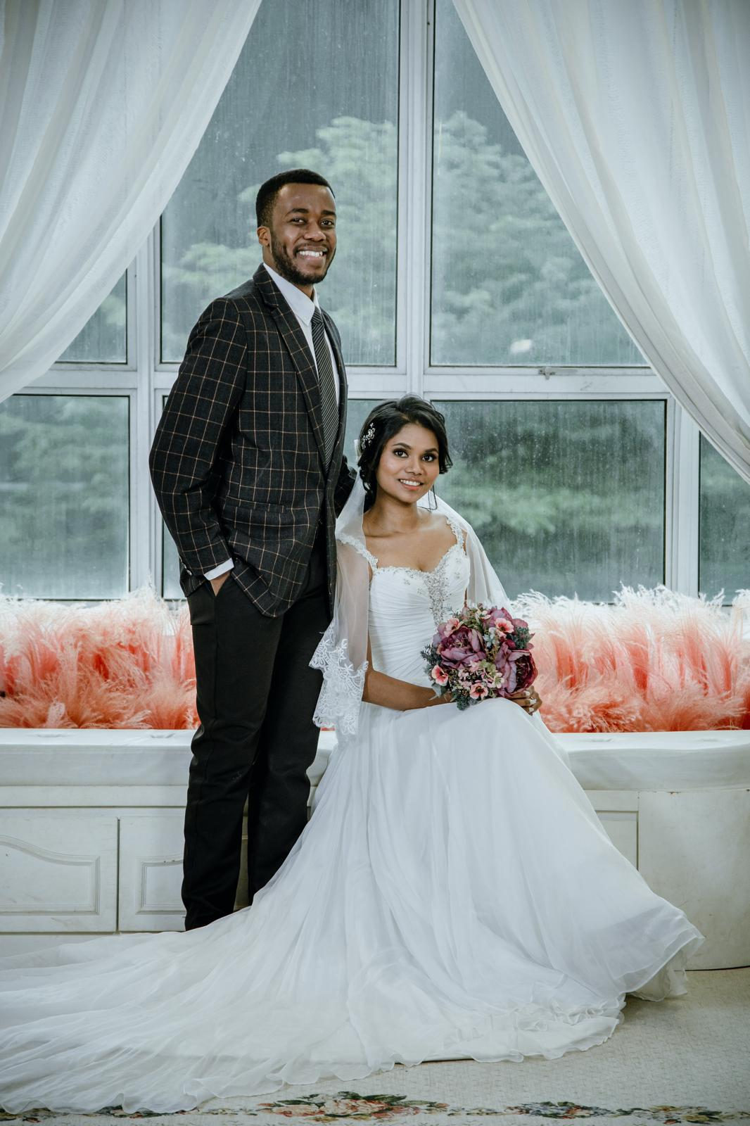 Aghalya in her wedding gown and Munshya in a tuxedo, standing by a bright window