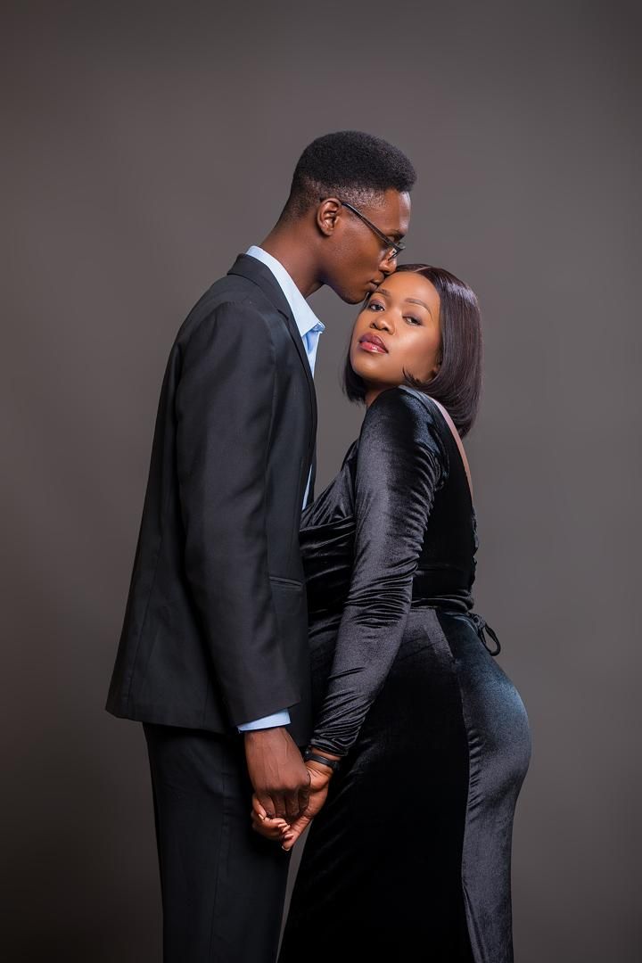 Elijah kissing Mwaka's forehead, both dressed in black against a dark studio backdrop