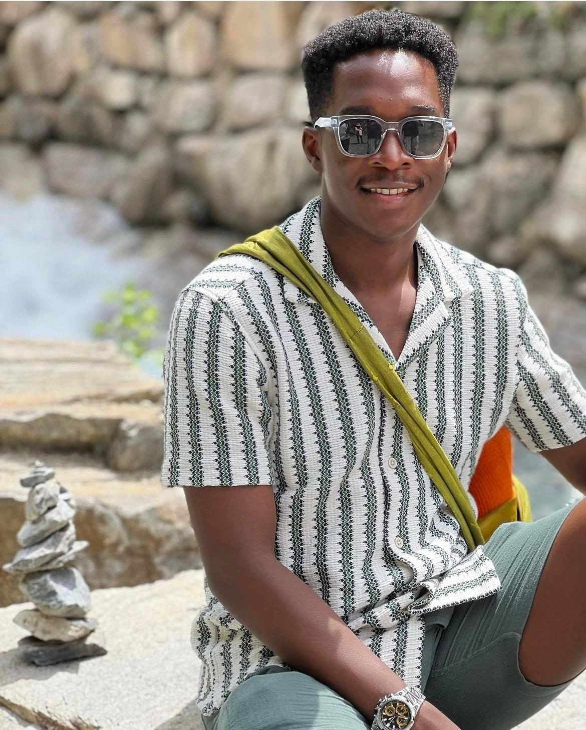 Mpaala in sunglasses and a patterned shirt, sitting on rocks by a stream