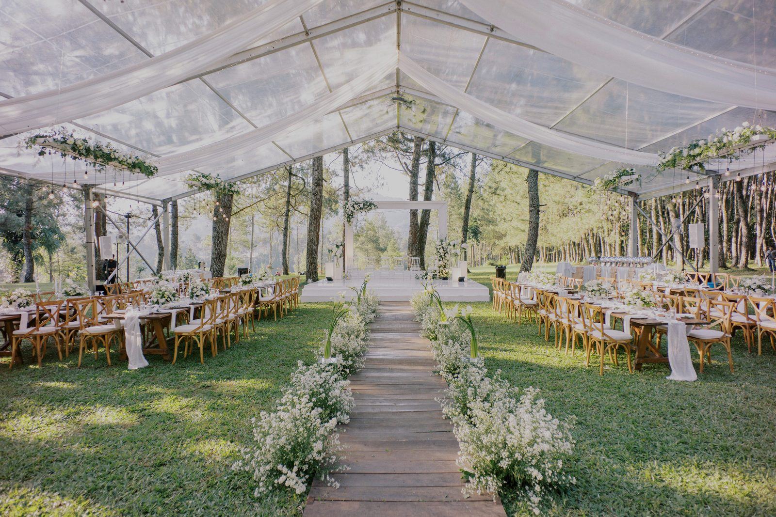 A clear wedding tent among pine trees, long tables set with white flowers along a wooden aisle